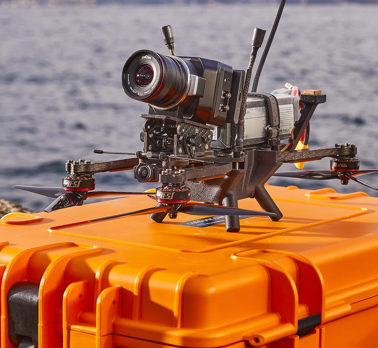 Heavy-lift FPV cinelifter drone carrying a compact cinema camera with a wide-angle lens, resting on a hard transport case beside water — Munich-based drone cinematography by NextFlow Studios — source: FARINS FRAMES